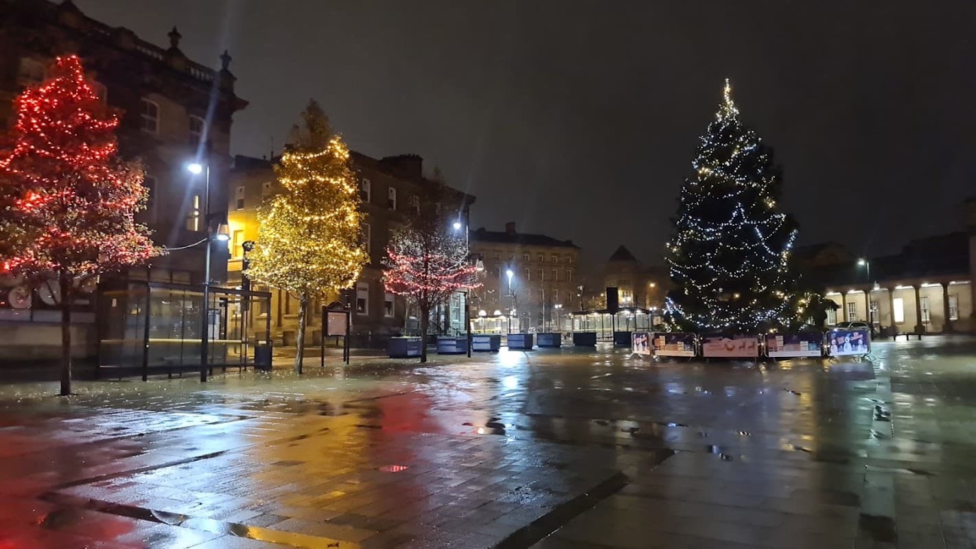 Festively lit trees and a decorated Christmas tree in a wet car park at night in Huddersfield, Yorkshire.