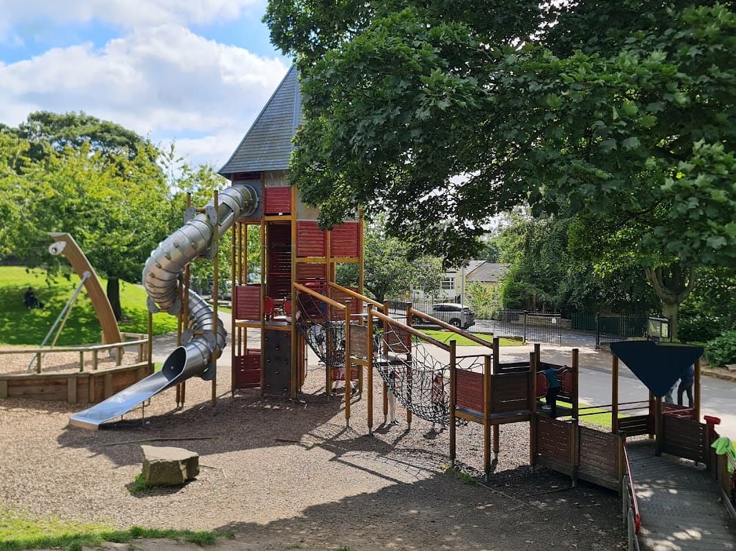 Playground equipment featuring a slide, climbing net, and a tower, surrounded by greenery and trees in Greenhead Park.