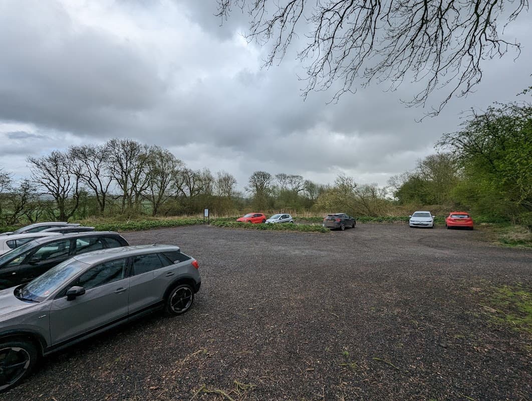 Gravel car park with several parked cars surrounded by trees and overcast skies in Huggate, Yorkshire.