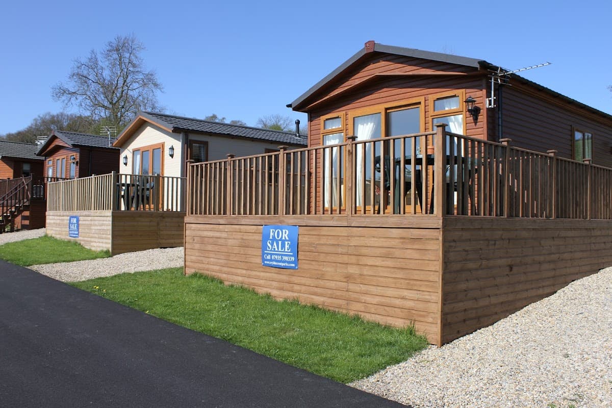 Two holiday cabins with wooden decks for sale, surrounded by gravel paths and greenery under a clear blue sky.
