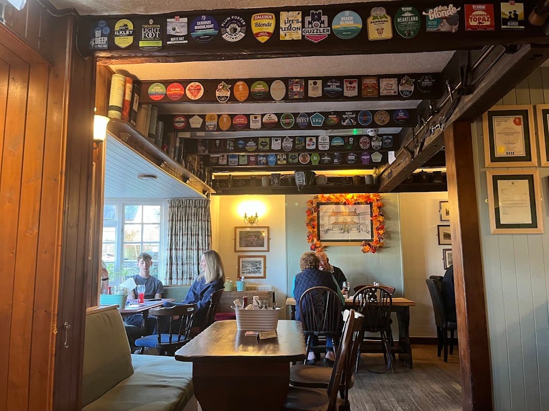 Cozy bar interior with wooden beams, patrons at tables, and a ceiling adorned with beer pump clips.