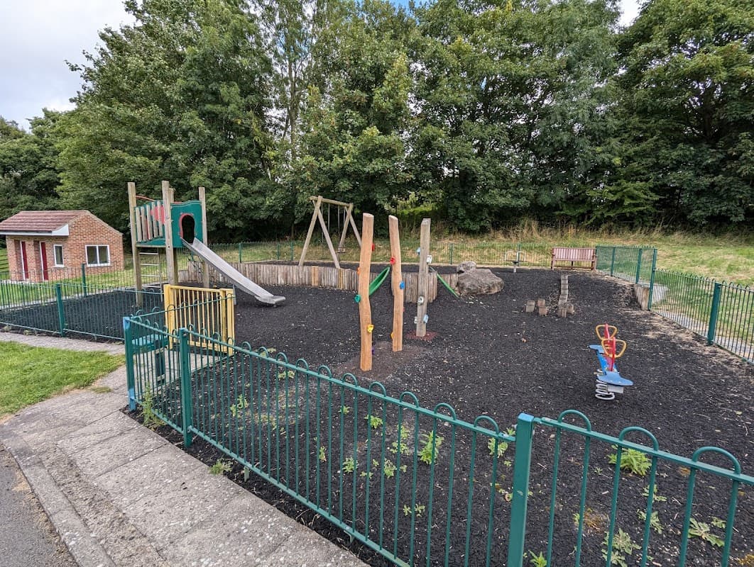 Playground with a slide, climbing structure, swings, and benches surrounded by a green fence and trees in the background.