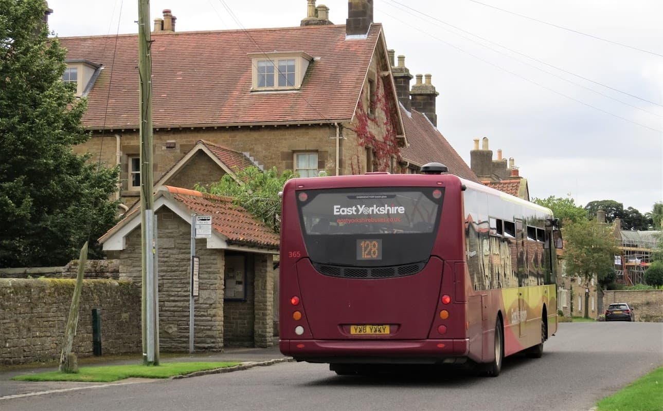 A bus labeled "East Yorkshire 128" drives past a stone building with a red roof in Hutton Buscel, Yorkshire.