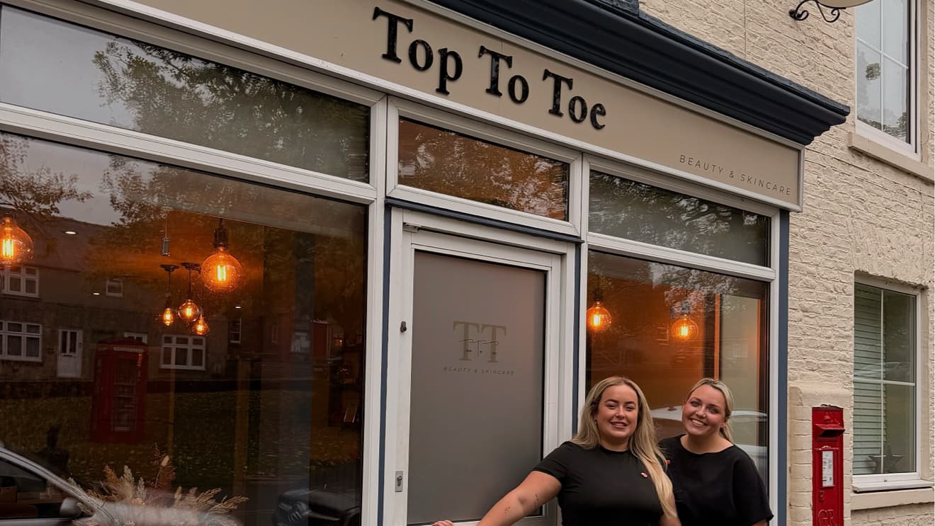 Two smiling women stand outside the "Top To Toe Beauty & Skincare" shop, featuring large windows and stylish signage.