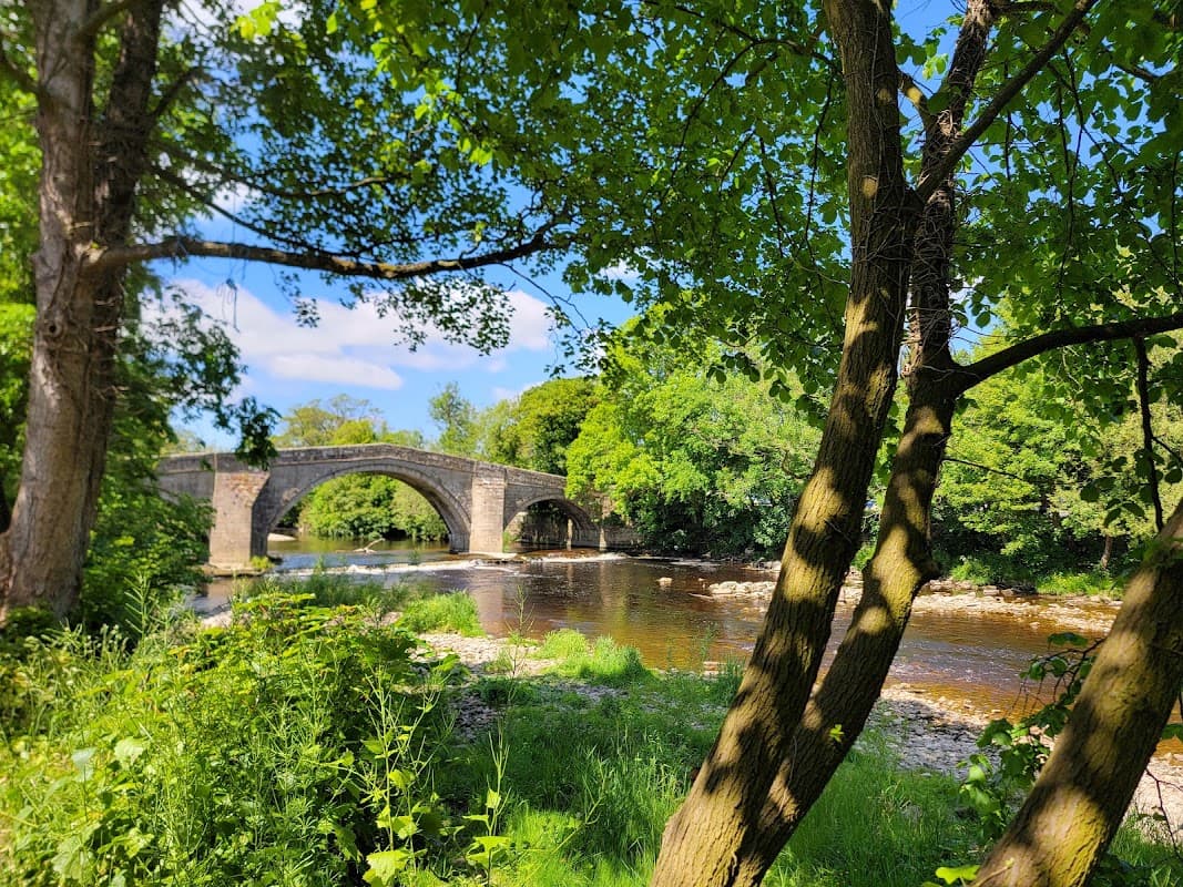 Stone bridge over a river, surrounded by lush greenery and trees under a bright blue sky.
