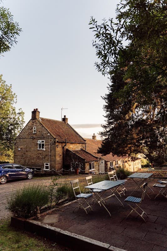 Charming stone guest house with a sloped roof, surrounded by trees and outdoor seating area in Ingleby Arncliffe, Yorkshire.
