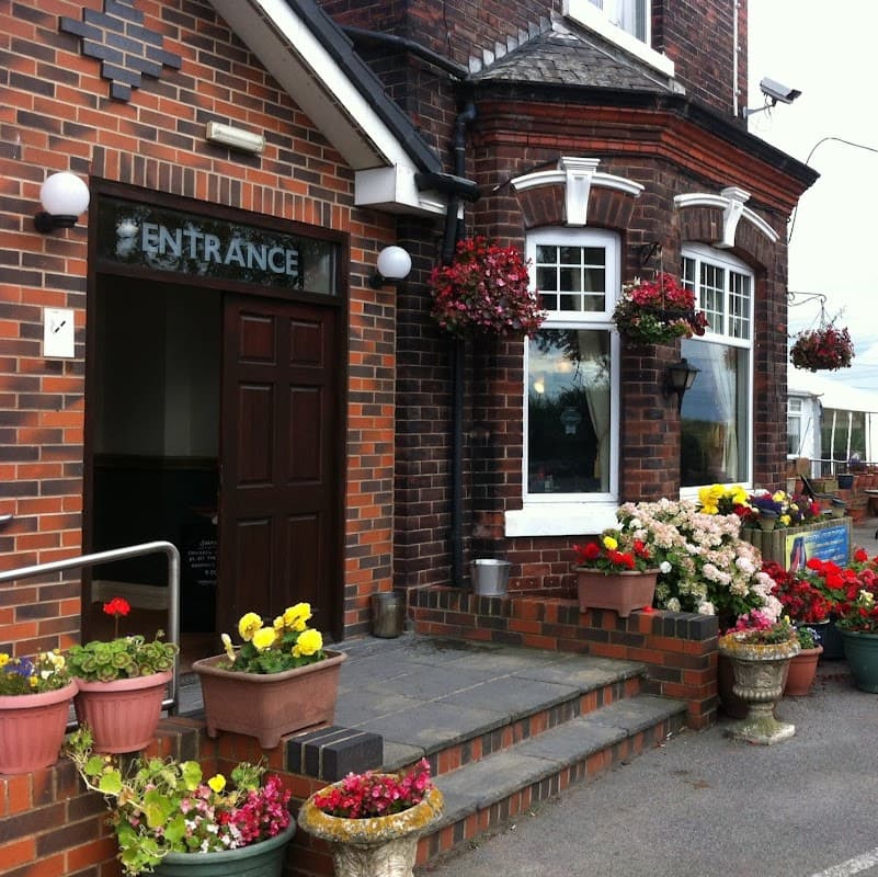 Brick building entrance with a sign, surrounded by colorful flower pots and hanging baskets.
