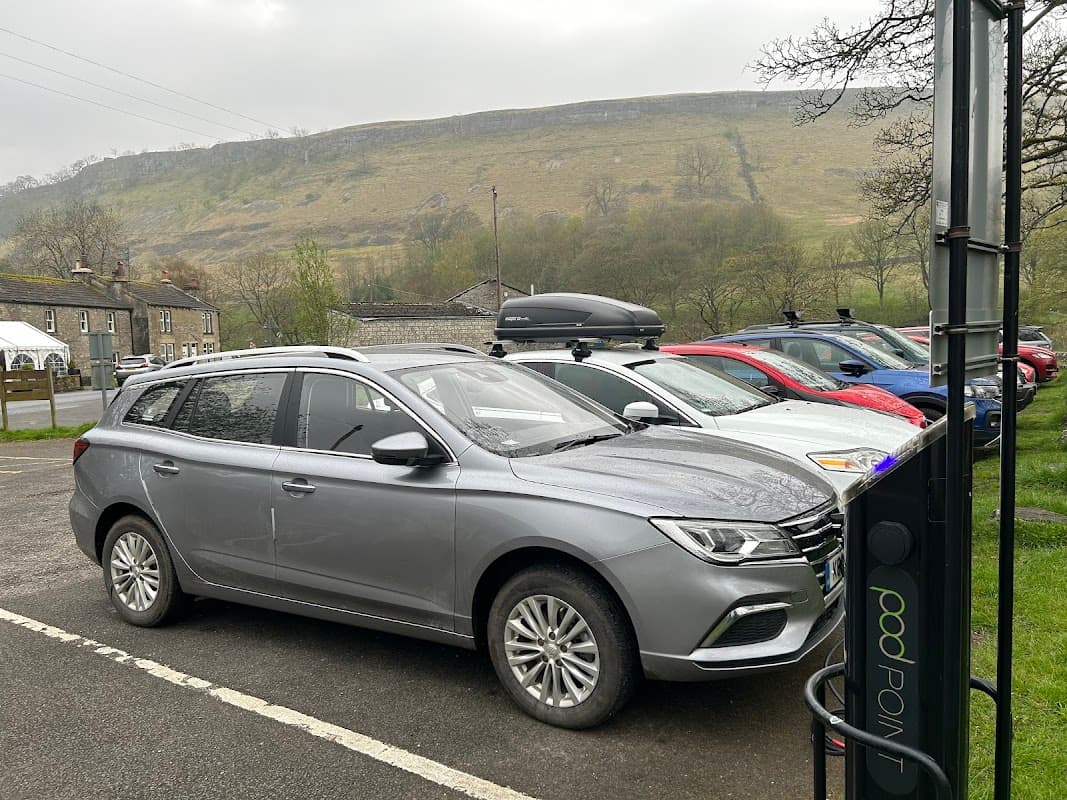 Pod Point charging station with a gray car parked nearby, surrounded by other vehicles and a hilly landscape in Kettlewell.
