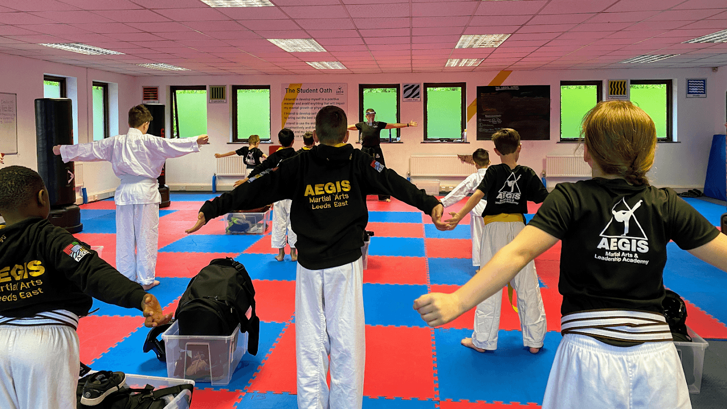 Martial arts students in black AEGIS uniforms practicing in a colorful dojo with mats and a trainer in the background.