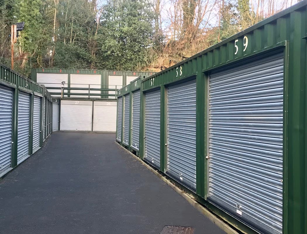 Green storage units with metal roll-up doors line a paved pathway, surrounded by trees in a quiet area.