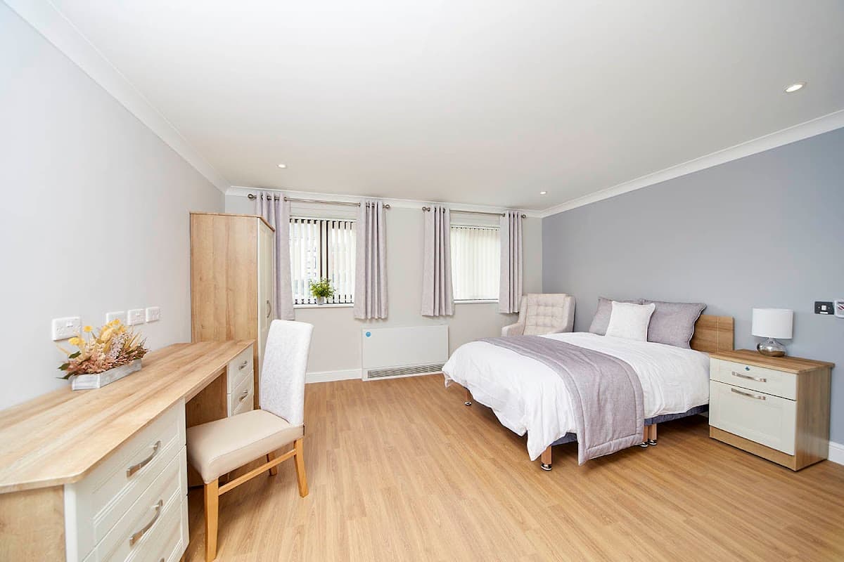 Bright, spacious bedroom with a bed, bedside table, dresser, and large windows, featuring light-colored walls and wooden flooring.