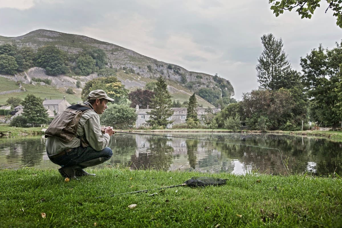 A man crouches by a pond, fishing, with lush greenery and limestone hills in the background.