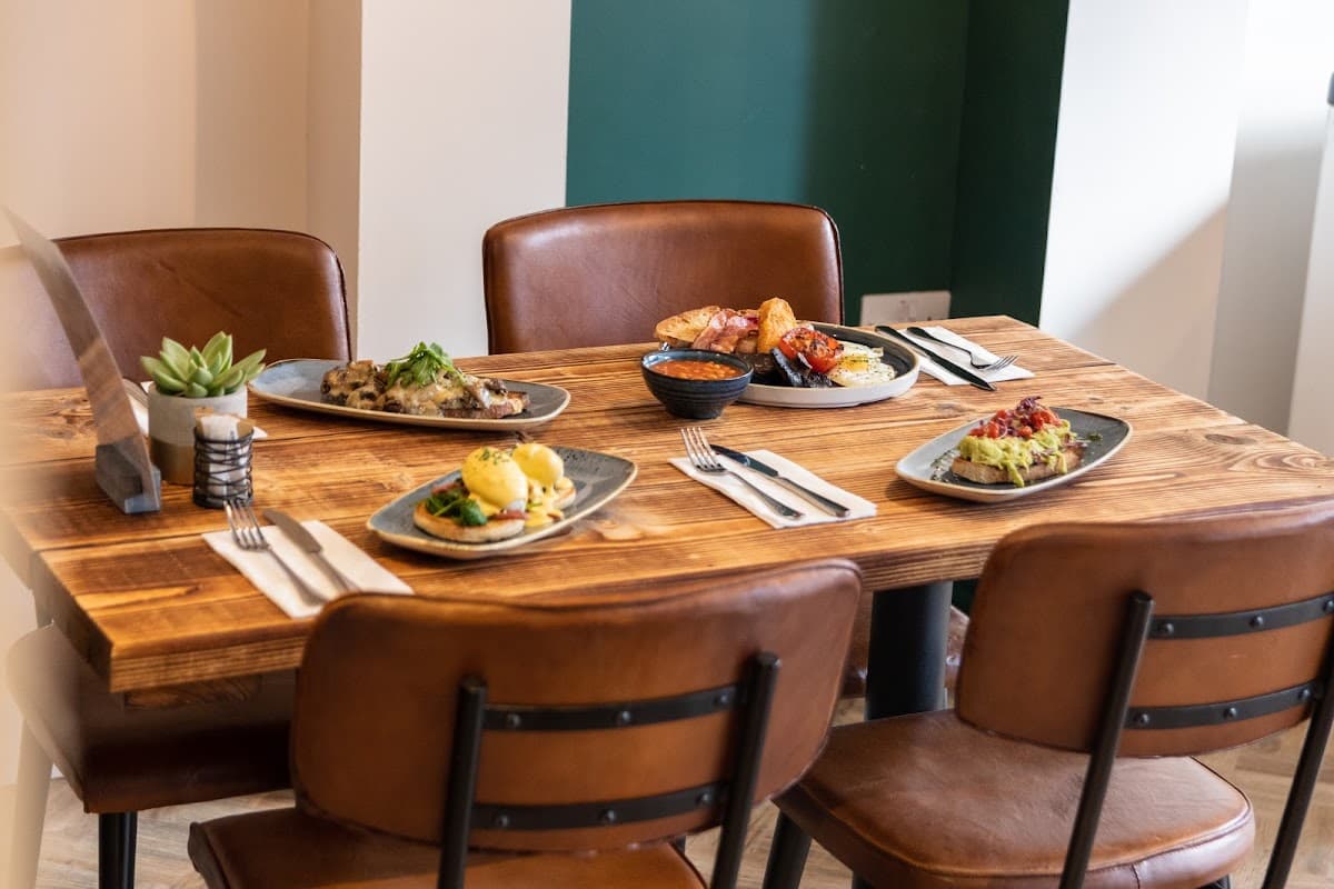 A wooden table set with five plates of food, cutlery, and a small plant in a pot, surrounded by leather chairs.