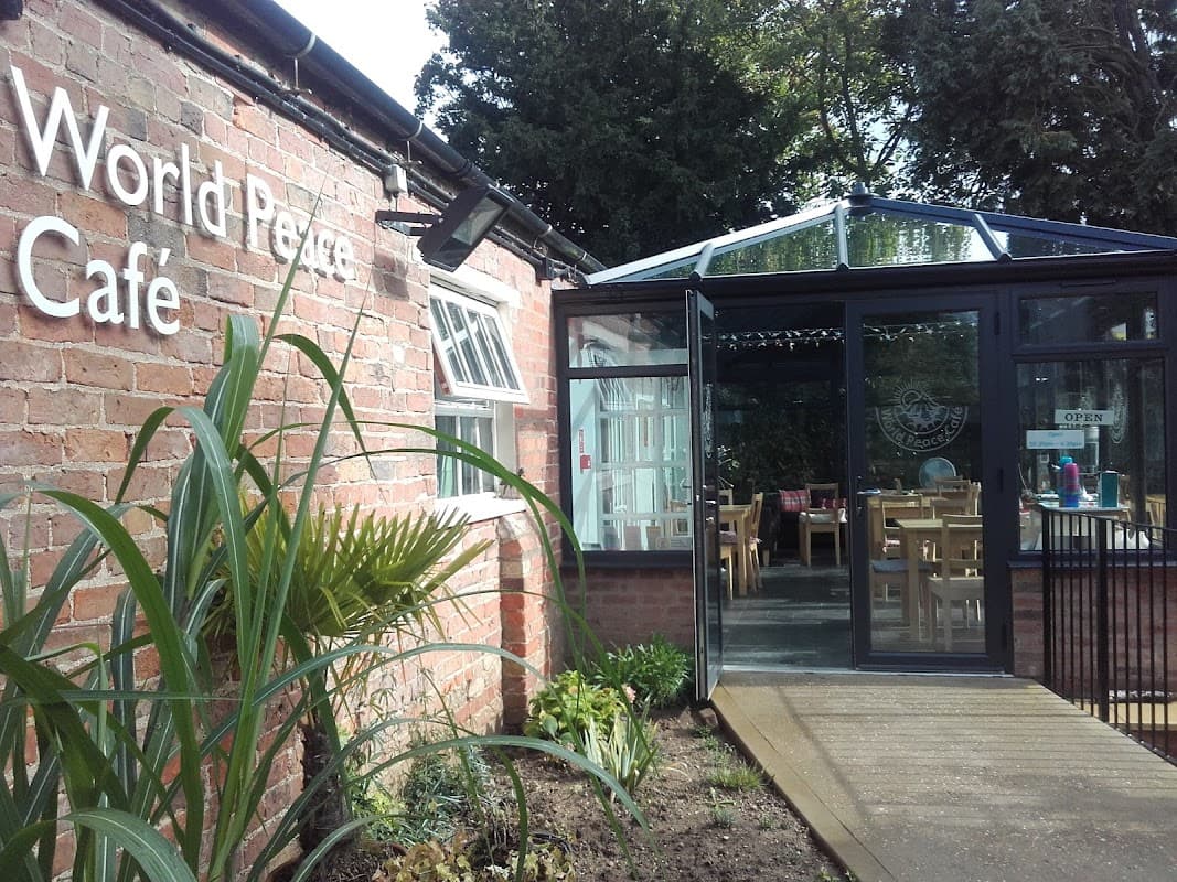 Entrance to World Peace Cafe with glass conservatory, surrounded by greenery and rustic brick walls.