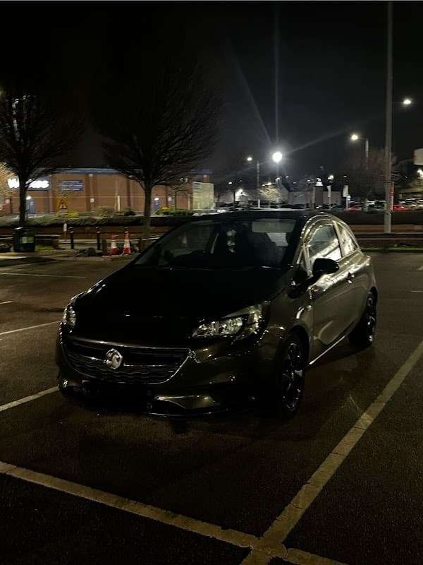 A black car parked in a lot at night, with streetlights and trees in the background.