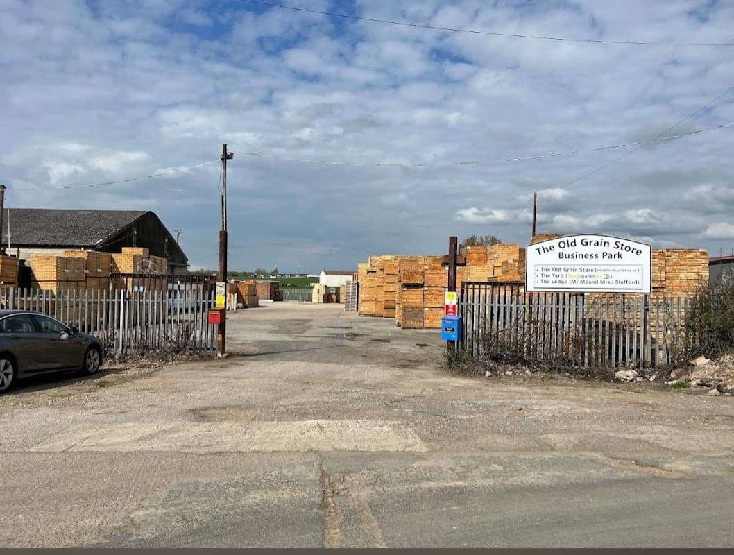Entrance to The Old Grain Store Business Park, with stacked wooden crates and a gravel road under a cloudy sky.