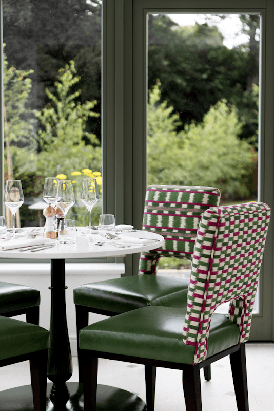 Elegant dining setup with a round table, green chairs, glassware, and yellow flowers, surrounded by lush greenery.