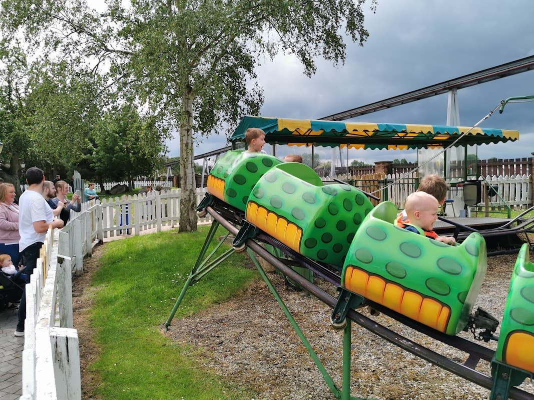 Children in green caterpillar-themed ride cars, with spectators nearby, under a cloudy sky at Flamingo Land attraction.