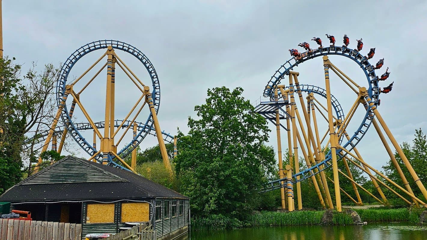 Roller coaster with yellow and blue tracks looping above a lake, surrounded by trees and a wooden building.
