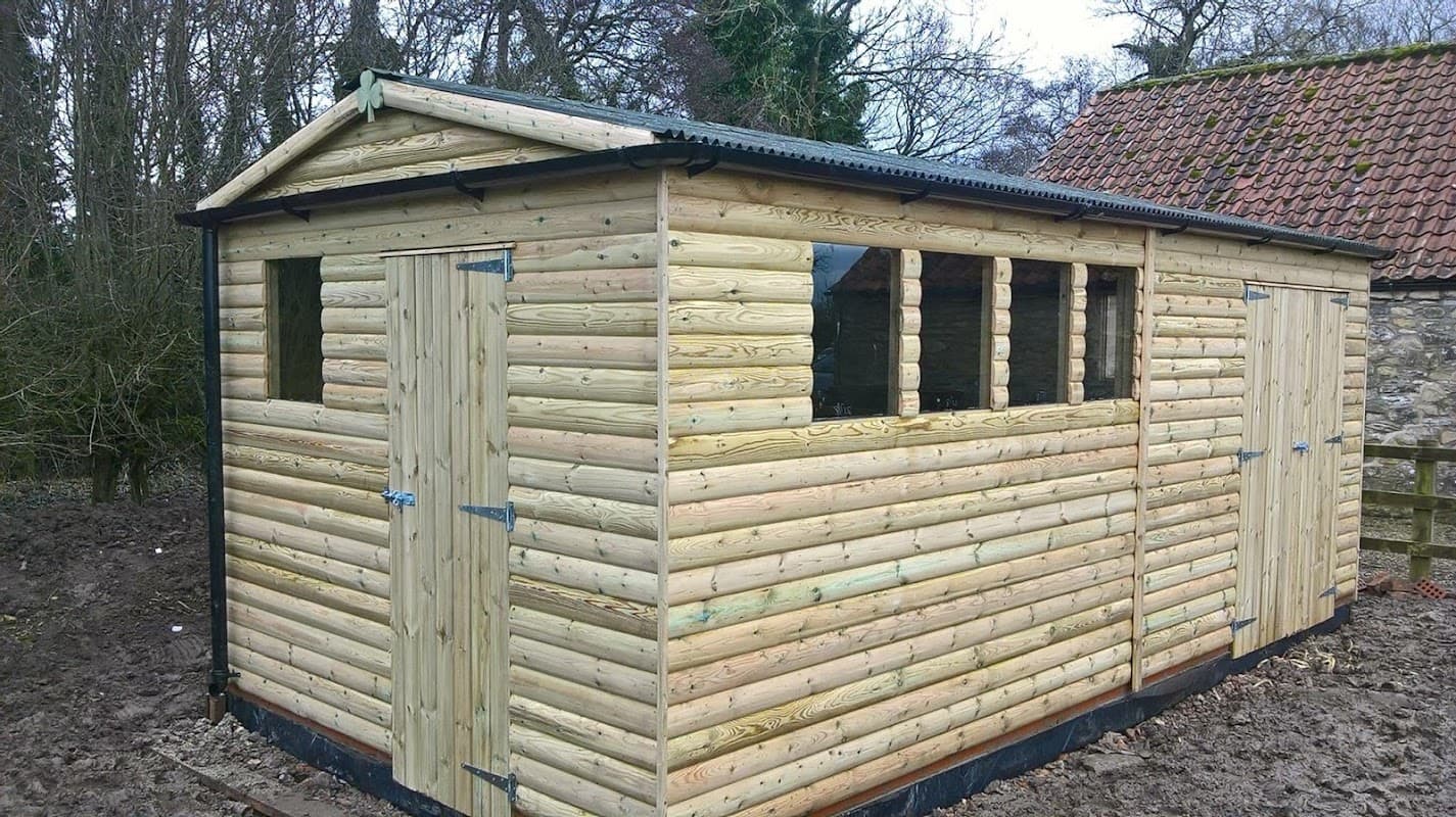 Wooden shed with a sloped roof, multiple windows, and double doors, set in a rural area with trees in the background.