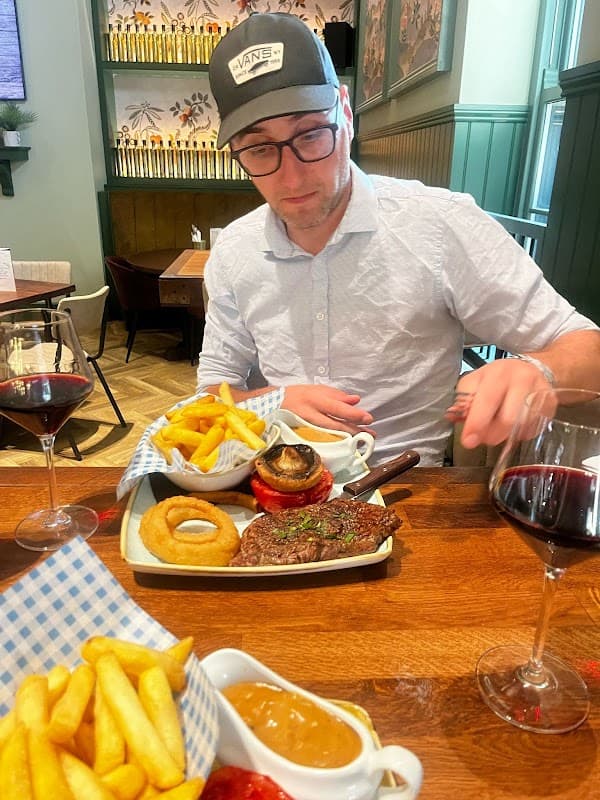 A man in a white shirt and glasses sits at a table with steak, fries, onion rings, and red wine in a cozy restaurant.