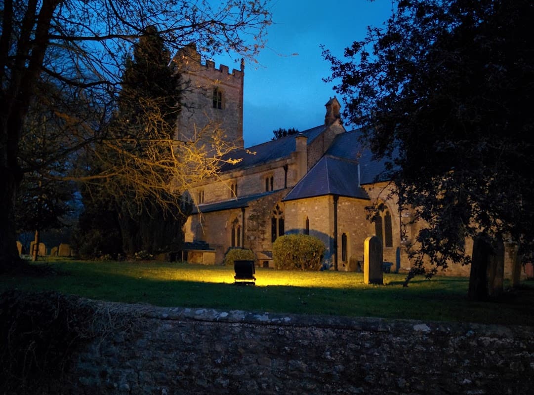 St Michael's church illuminated at dusk, surrounded by trees and gravestones in Kirklington, North Yorkshire.