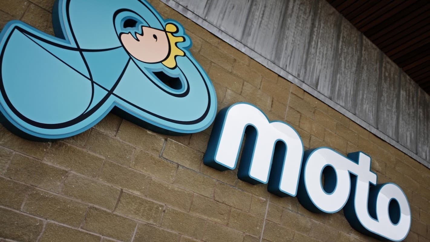 Bright blue "moto" logo with a cartoon character, displayed on a textured stone wall at Moto Ferrybridge.