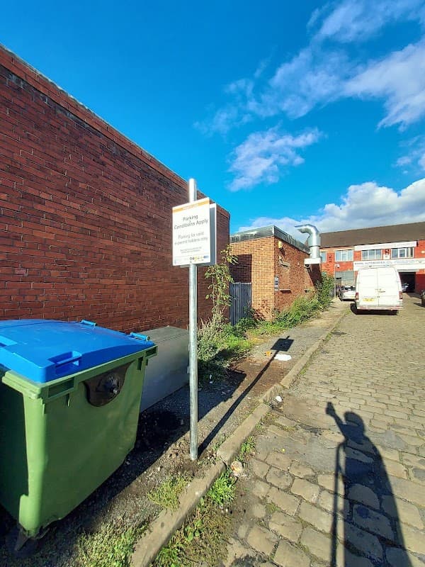 Sign indicating parking rules next to a green bin, with a van parked on a cobblestone path under a blue sky.