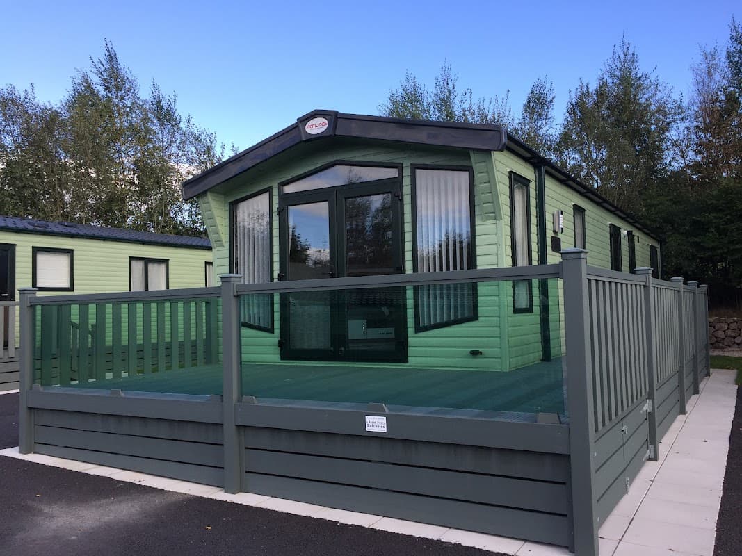 Green caravan with a deck, surrounded by trees, in Langcliffe Caravan Park, Yorkshire. Clear blue sky above.