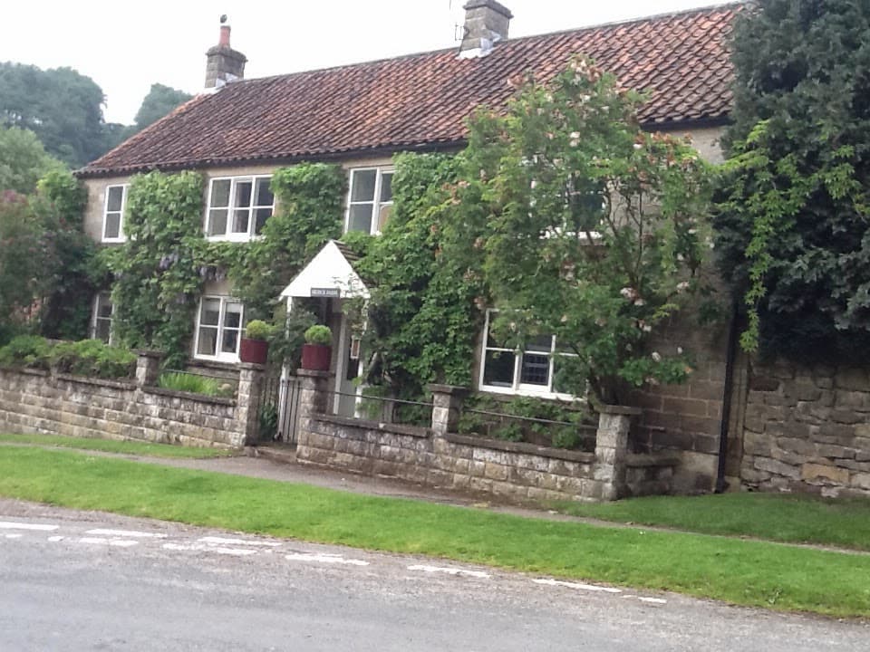 Charming stone cottages with a red-tiled roof, surrounded by greenery and a stone wall, in a rural setting.