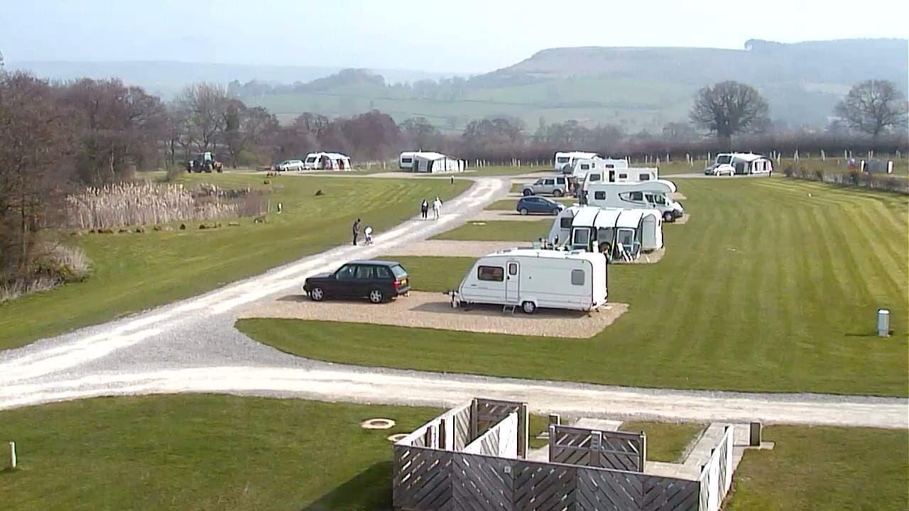 Caravans and vehicles parked along a gravel path in a green, grassy hillside setting with trees and distant hills.