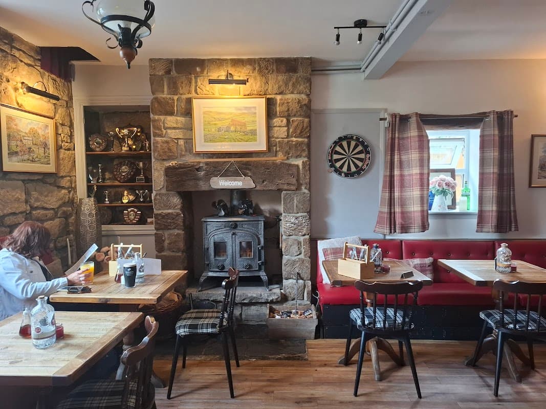 Cozy pub interior with stone walls, wooden tables, a dartboard, and a welcoming fireplace. Warm lighting enhances the atmosphere.