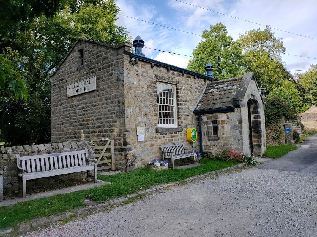 Stone building with a sign reading "Village Hall for Hire," surrounded by trees and benches, in Leathley, Yorkshire.