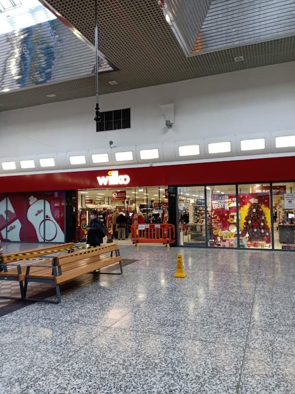 Entrance to Wilko store with festive decorations, a bench, and caution sign on the floor in a shopping center.