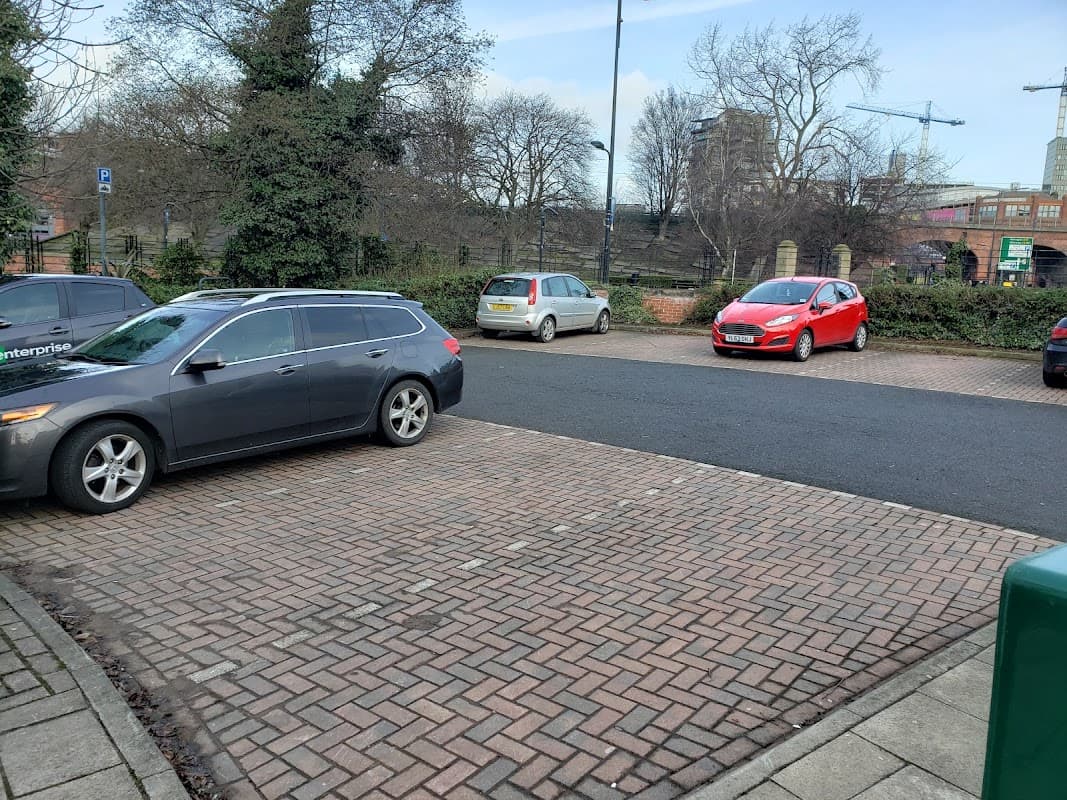 Maude Street Parking Area in Leeds, featuring parked cars and a paved surface, with trees and buildings in the background.