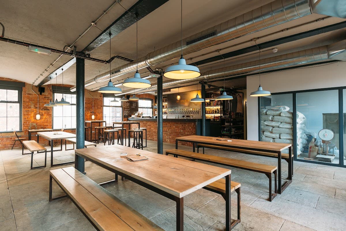Spacious interior with wooden tables, benches, industrial lighting, and a bar area featuring bottles and brewing equipment.
