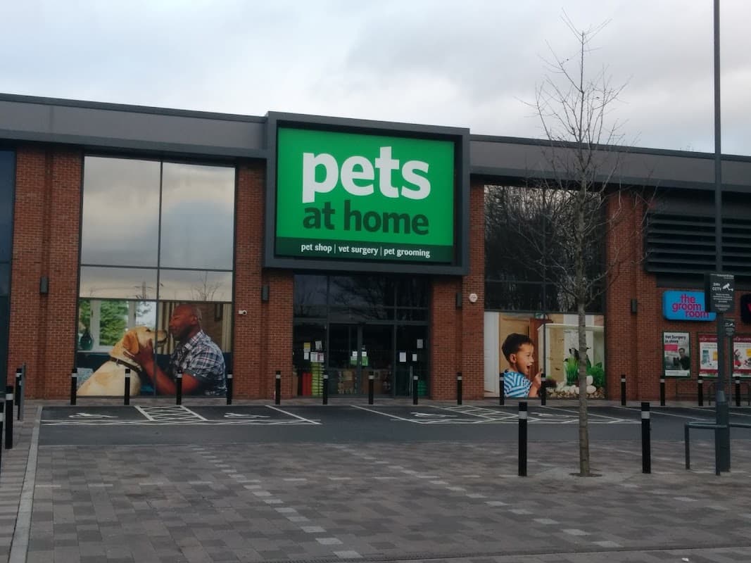 Bright green sign reading "pets at home" on a brick storefront with images of people and pets. Parking area visible.