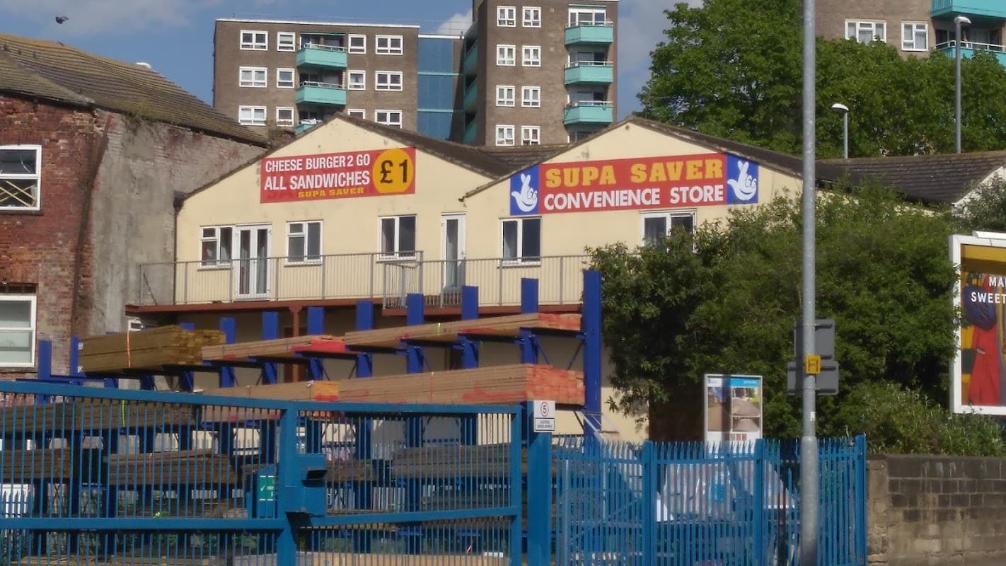 Supa Saver convenience store with signage for burgers, surrounded by buildings and blue fencing in Leeds.