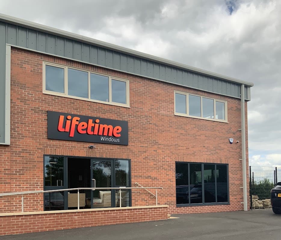 Brick building with large windows, "Lifetime Windows" sign, and a paved area in front. Cloudy sky in the background.