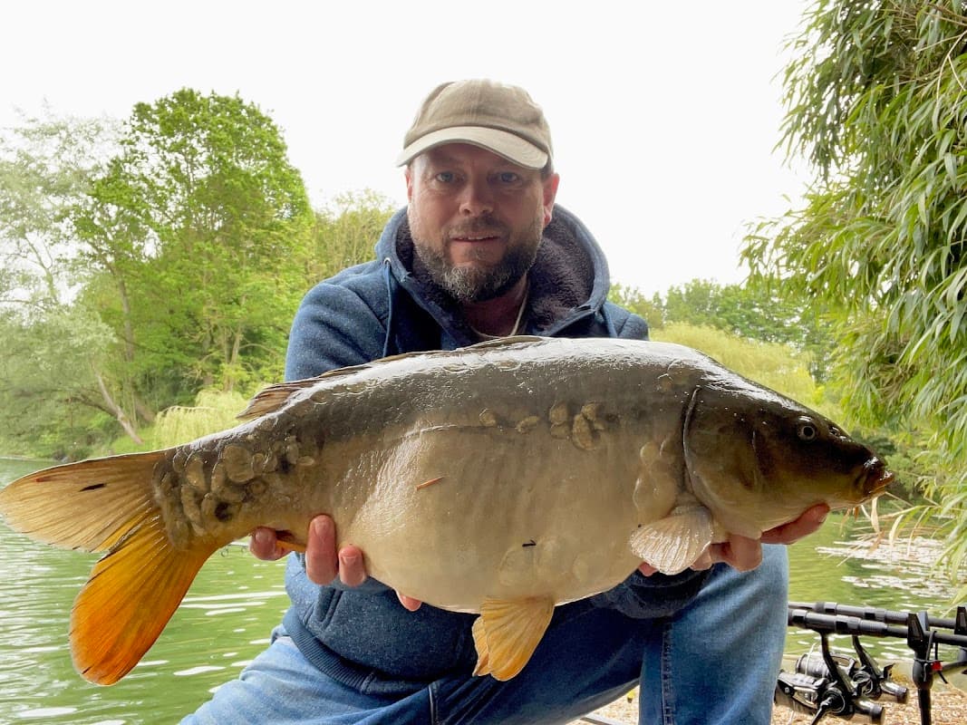 A man holding a large fish by a lake, surrounded by greenery and fishing rods in the background.