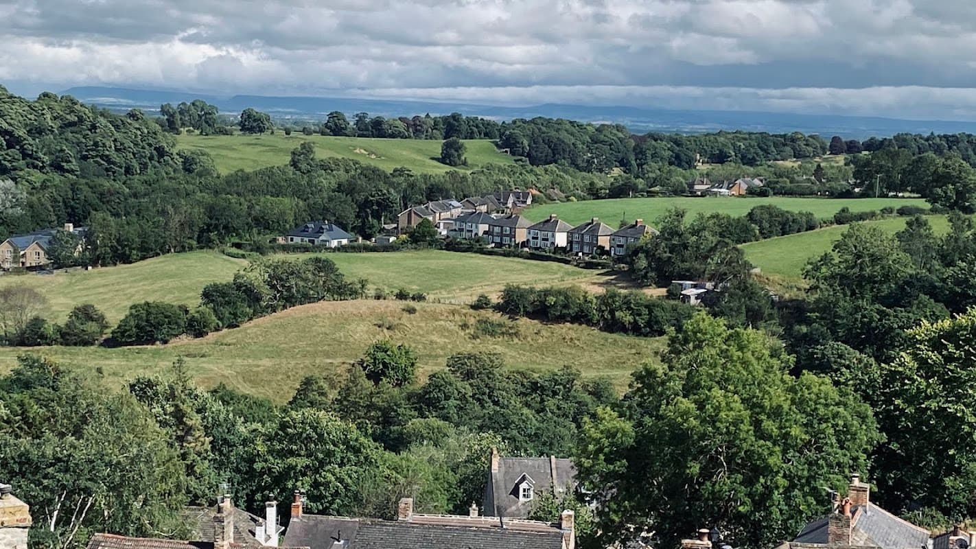 Leyburn Caravan Park nestled among lush green hills and trees, with houses visible in the distance under a cloudy sky.
