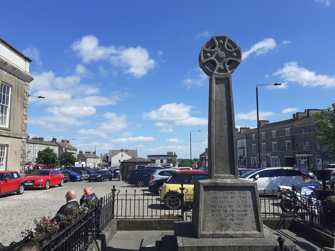 Celtic cross monument in a market square with parked cars and a blue sky in Leyburn, Yorkshire.