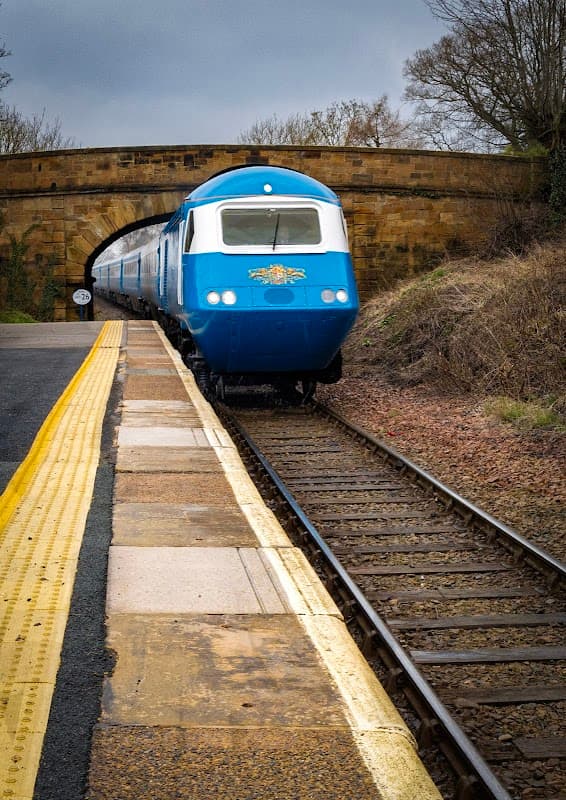 A blue train approaches a stone bridge at a railway station with a yellow platform edge and trees in the background.