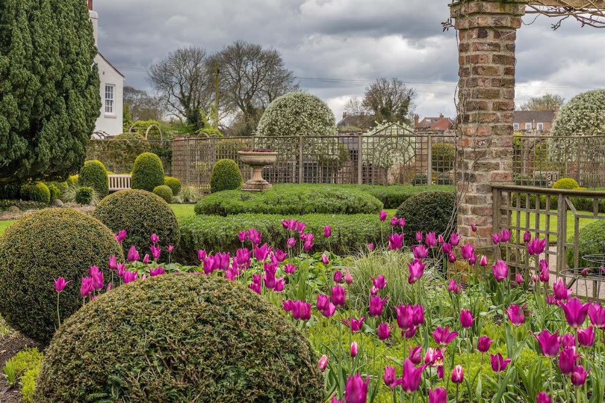 Lush gardens featuring rounded hedges, vibrant pink tulips, and a stone pillar against a cloudy sky in Littlethorpe.