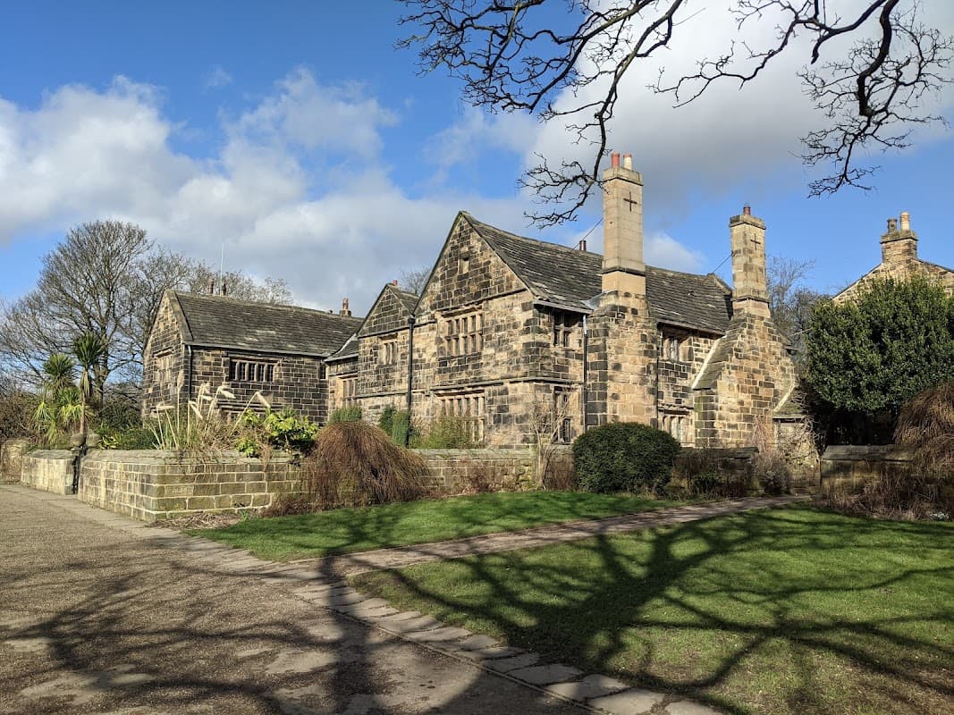 Historic stone building surrounded by lush greenery and trees under a partly cloudy sky. Pathway leads to the entrance.