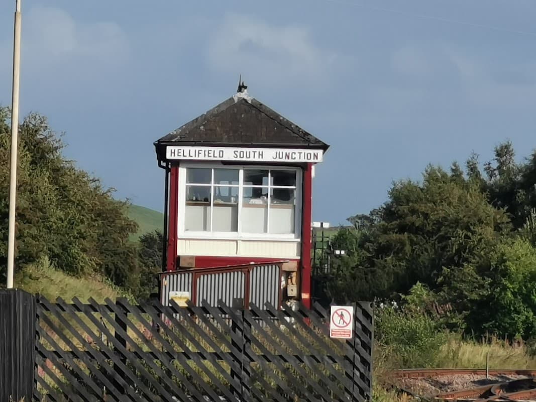 Hellifield South Junction signal box with a black wooden fence and green hills in the background under a clear sky.