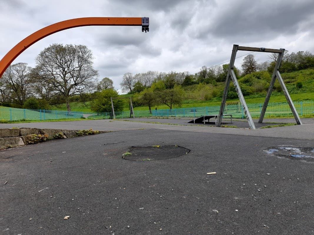 Empty car park with a swing set and a large orange structure, surrounded by green hills and cloudy skies.