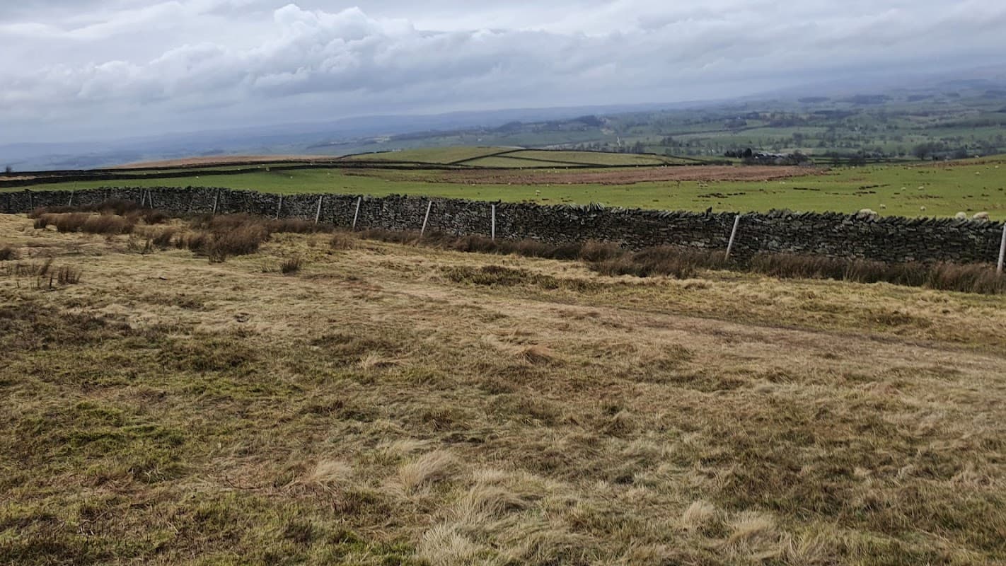 Open grassy area with a stone wall, rolling hills in the background, and cloudy skies over Lothersdale, Yorkshire.