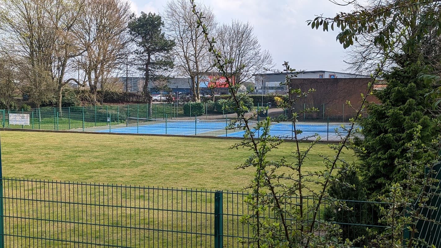 View of grassy area with tennis courts, surrounded by trees and fencing, under a cloudy sky in Coronation Park, Maltby.