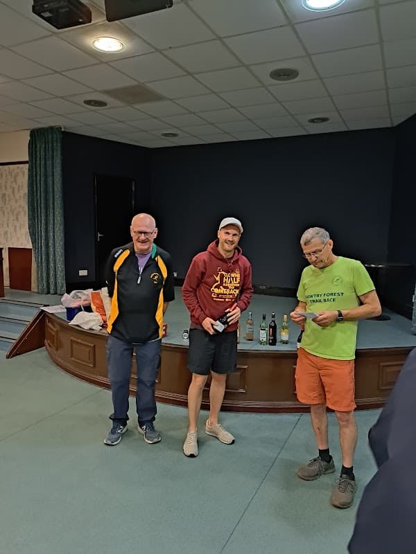 Three men stand in a bar setting, with a small table displaying bottles behind them. The room has dark walls and carpet.